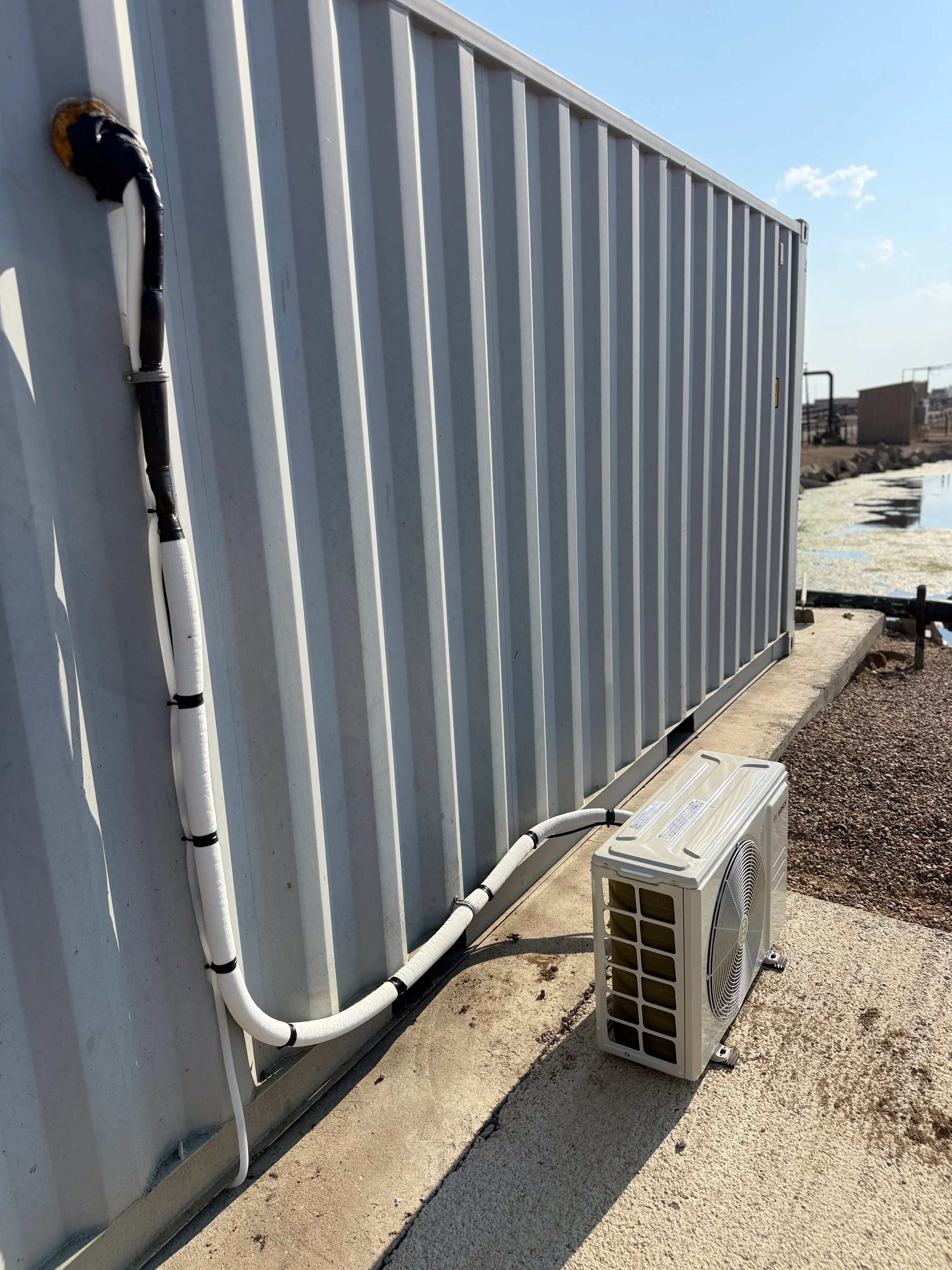 Ductless mini-split heat pump unit installed at base of white corrugated metal building with routed refrigerant lines