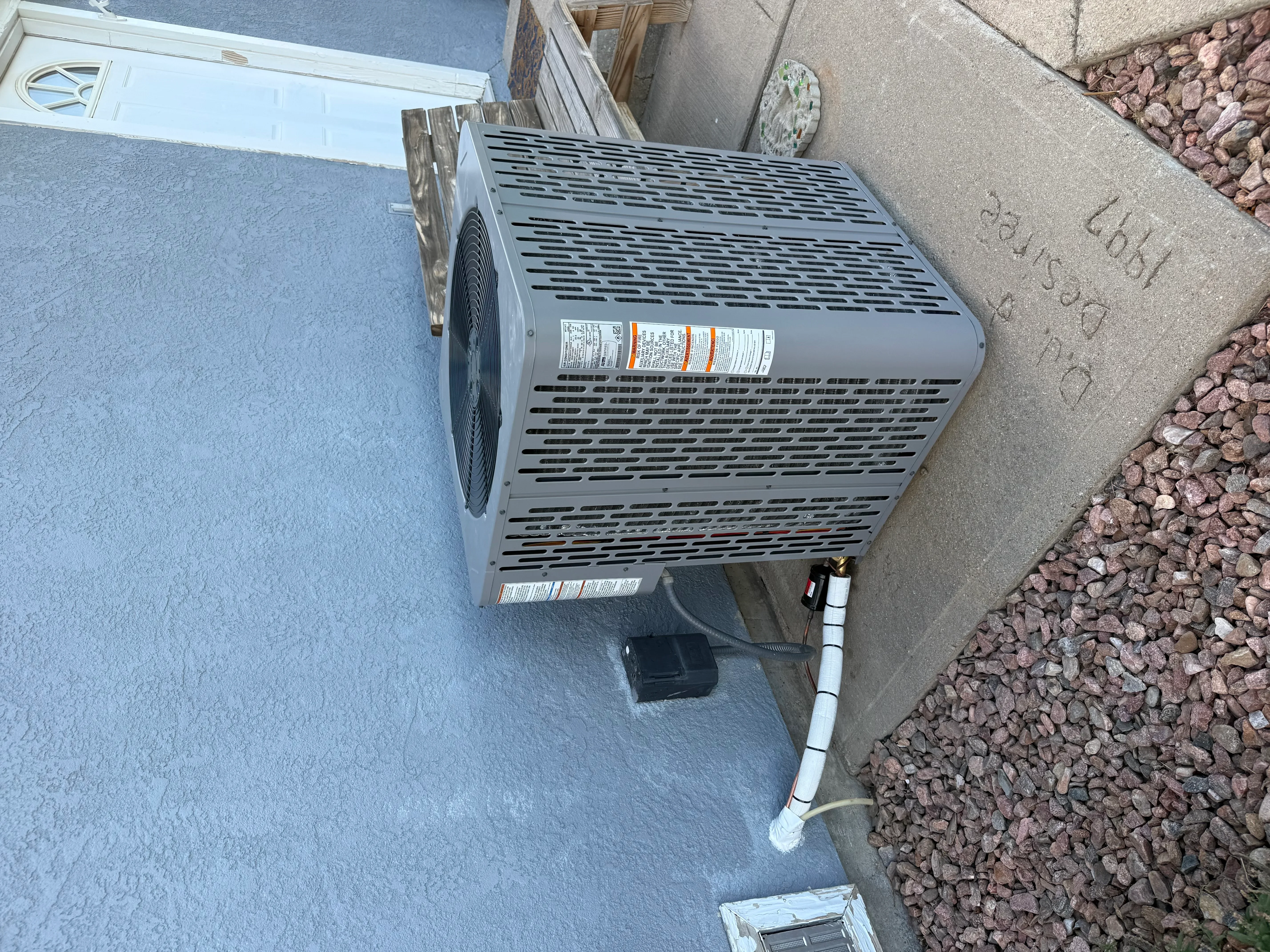 Gray AC condenser unit installed on concrete pad with white PVC drain line and red rock landscaping